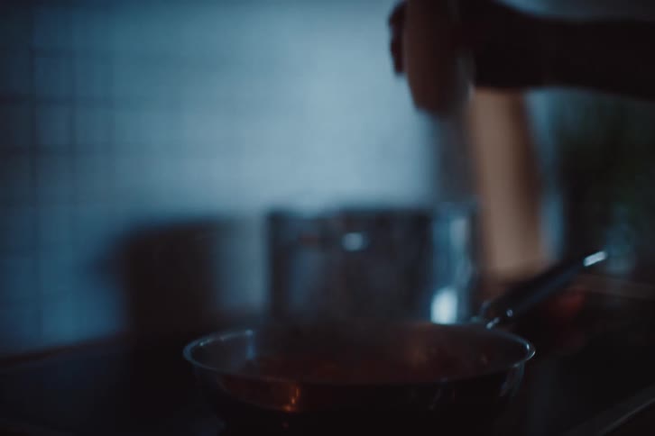 A hand pours liquid into a pan in a dimly lit kitchen setting