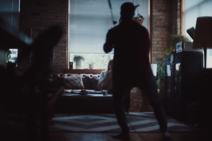 A person standing is holding a vacuum cleaner and dancing towards a person sitting on a sofa