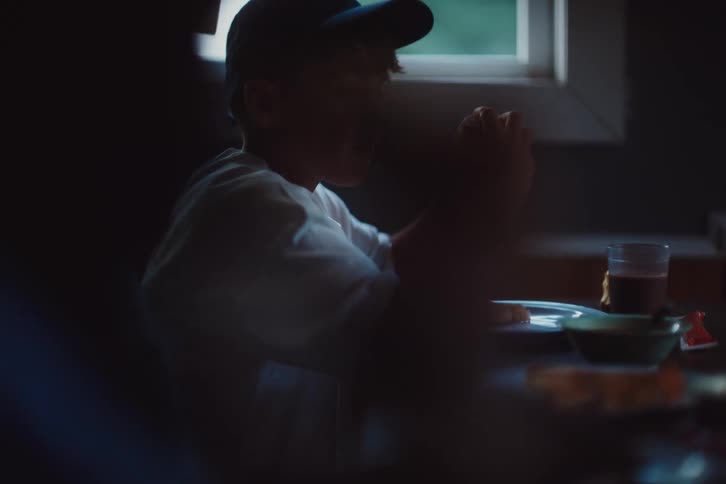 A little boy in a blue cap is sitting in front of a table and eating a sandwich and to the left of it there is a little girl who is taking something from the table