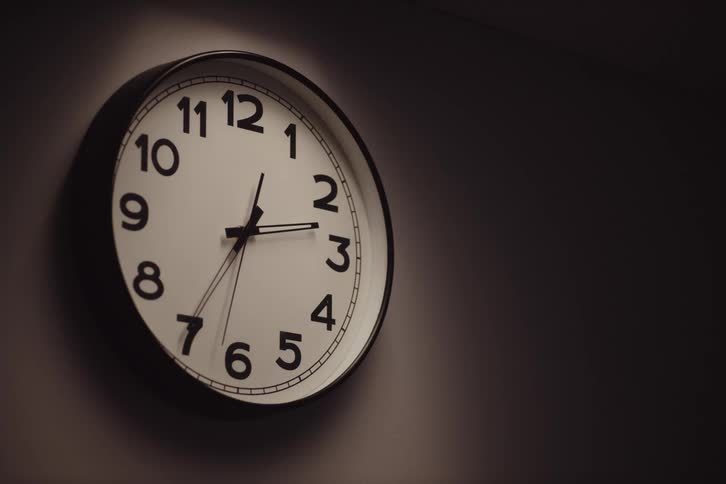 A large round wall clock hangs on a dark wall its hands pointing to three