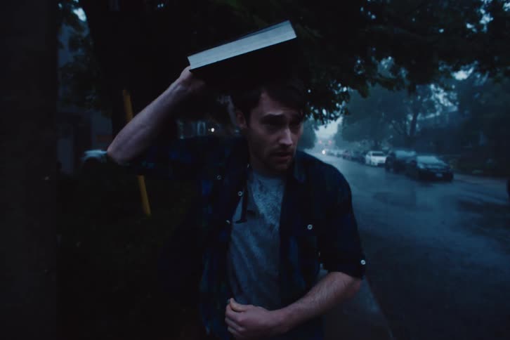 A young man holds a book over his head while walking in a suburb in the rain