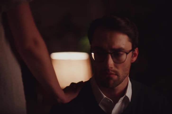 The face of a young man with glasses staring straight ahead and he has a depressed expression and then a person walks in front of him