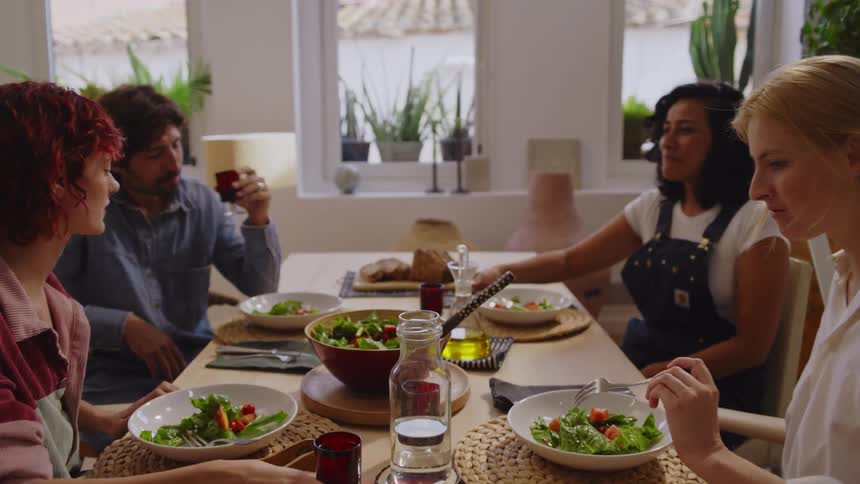 Four people sitting around a wooden dining table sharing a meal in a bright and cozy room with potted plants on the windowsill and warm natural light streaming in