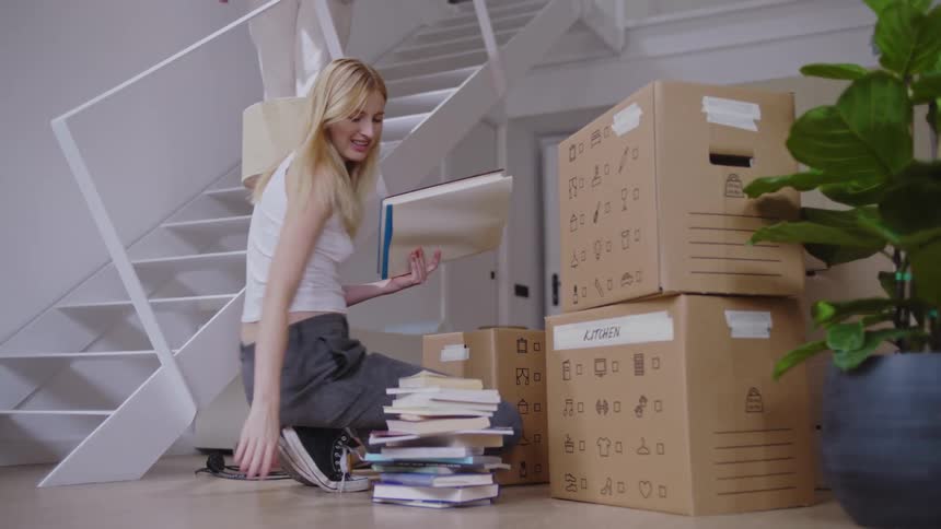 Woman walking down a staircase crouches to interact with a blonde woman unpacking a box of books