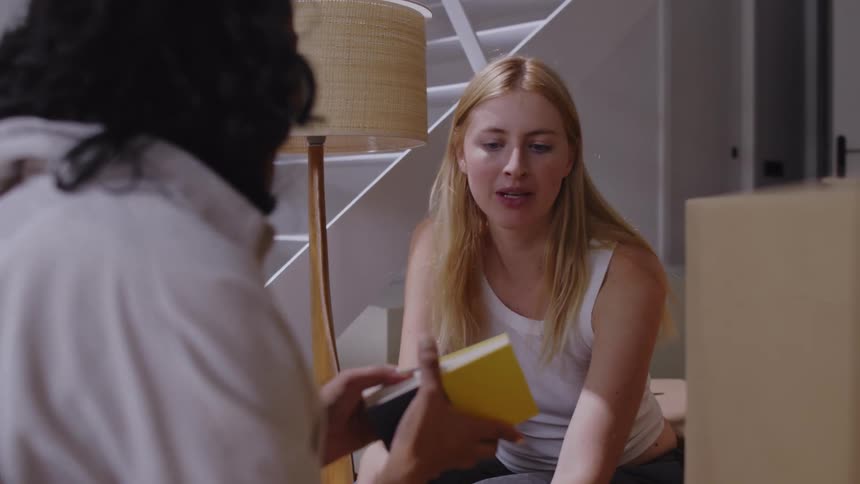 Women crouch in their home near large cardboard boxes and discuss a book