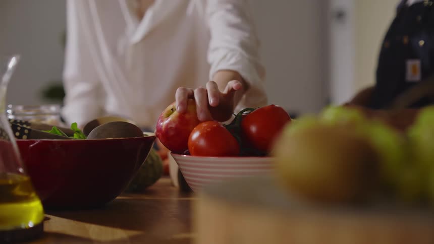 Table with fruits and vegetables and two women cooking in the background