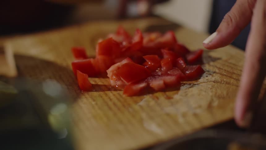 A persons hands are seen chopping a tomato on a wooden cutting board