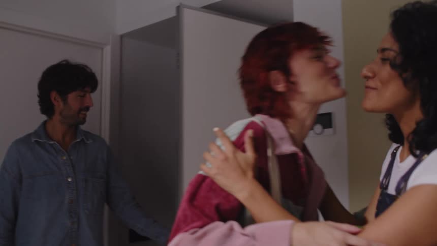 A woman smiles as she enters a room greeted by a hug from another woman with a man following behind her who is greeted by another woman