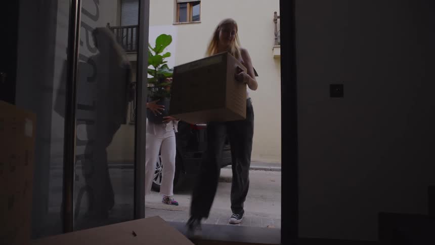 A couple is unloading cardboard boxes and potted plants from a vehicle parked near the entrance of an apartment building