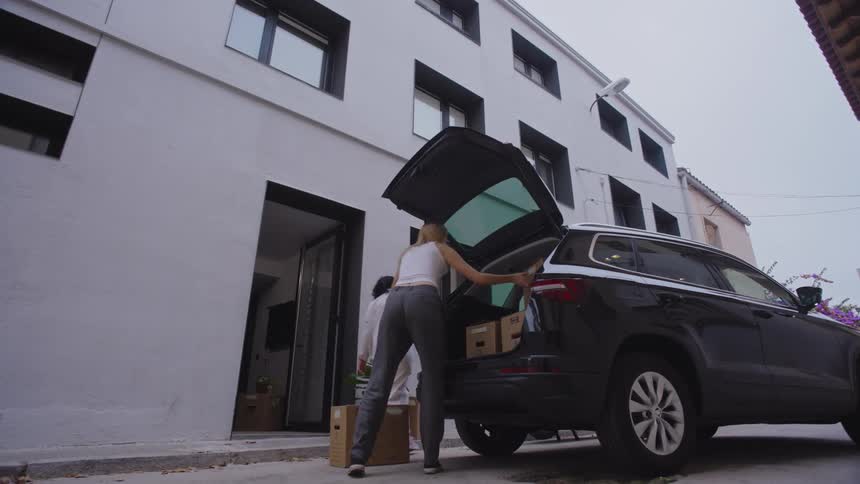 Women unload boxes from a car and carry items into a building during moving day