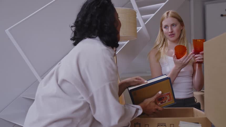 Two women one with a book and the other holding two glasses are having fun while standing in a living room surrounded by boxes
