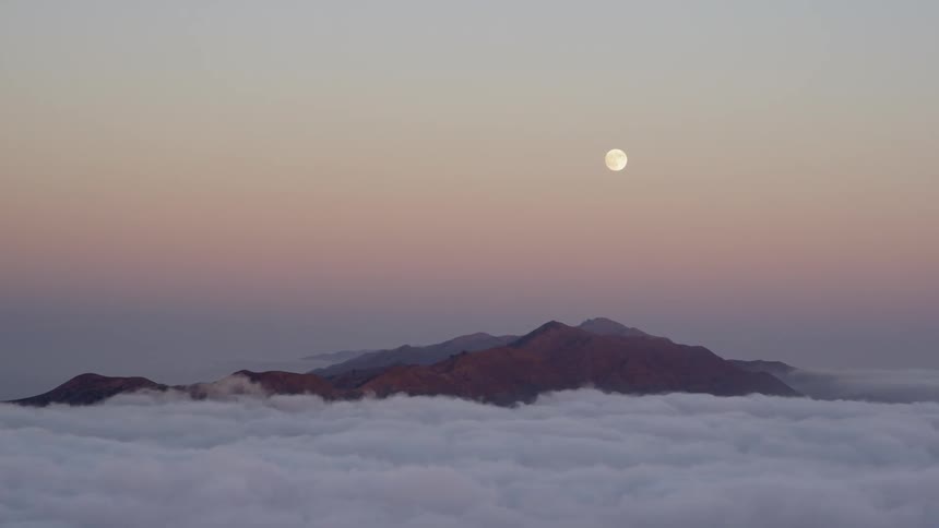Mountaintops protruding through clouds with a full moon in the sky