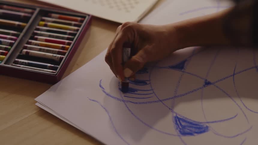 A hand colors in abstract shapes with blue crayon on paper next to a box of colorful crayons