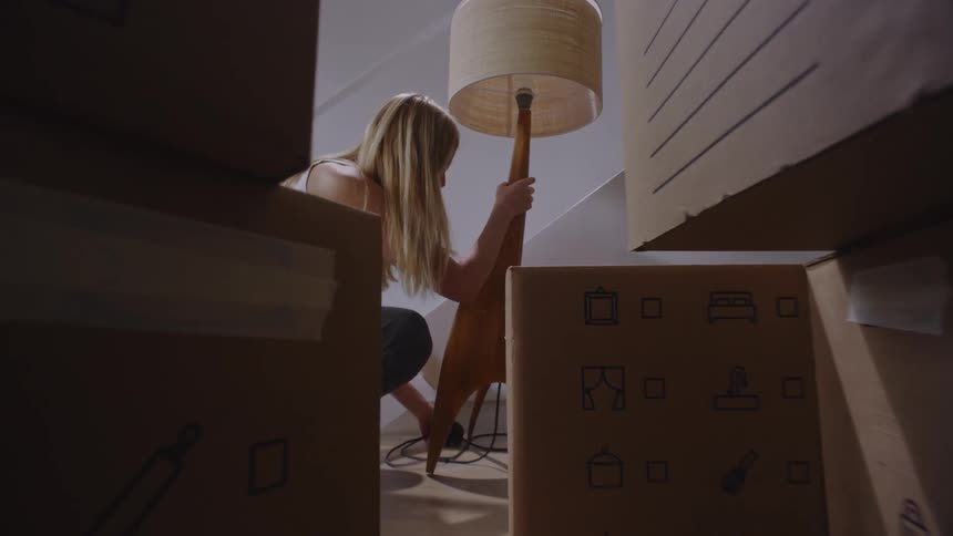 A blonde woman picks up a lamp near stacked moving boxes in a room