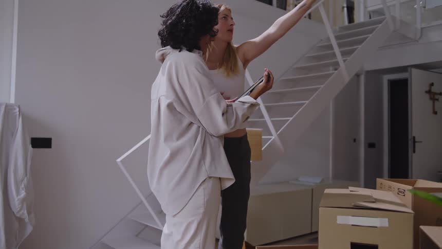 Two women holding hands in a living room talking and one of them hands the other a book