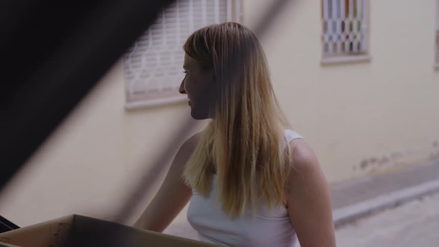 A blonde woman smiles at another woman passing by while she removes a box from her vehicle