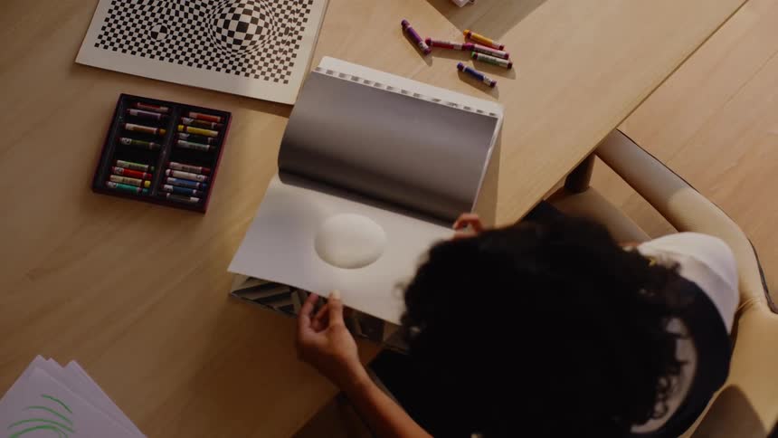A table with a sketchbook featuring abstract designs surrounded by colorful crayons a box of art supplies and geometric patterns on nearby sheets of paper