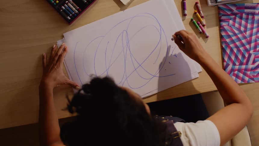 A person is drawing large abstract lines with a blue crayon on a white sheet of paper at a desk filled with colorful art supplies