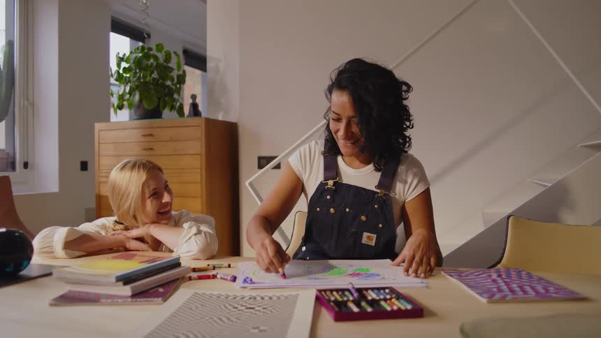 Woman crouches down next to another woman drawing in color and talks to her