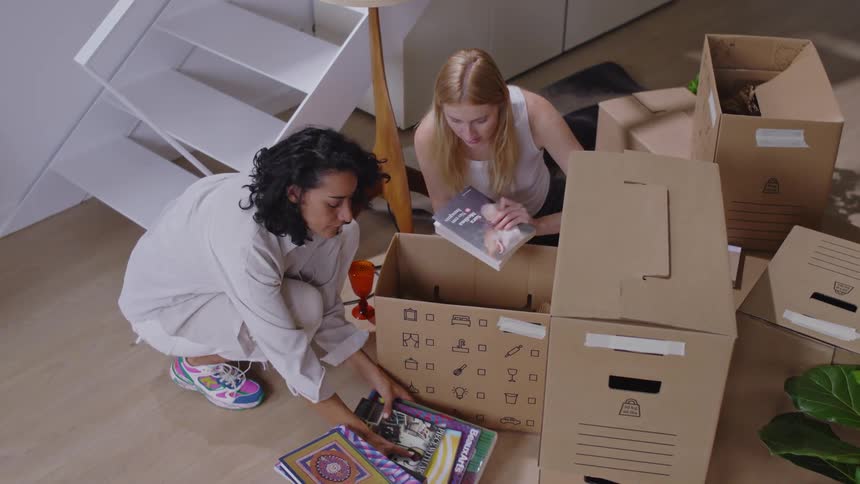 Two women chat while looking at books taken out of a moving box