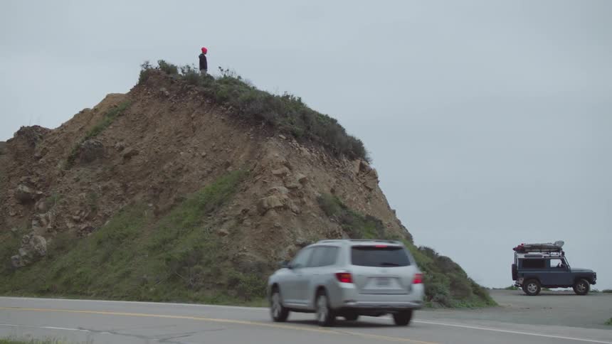 A person stands on top of a tall rock near a blue jeep at the beach