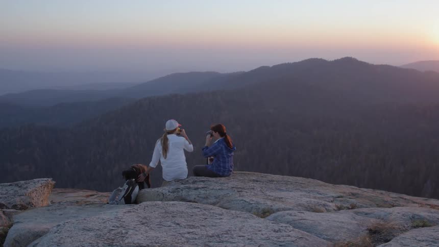 Two women and a dog are sitting on a cliff's edge enjoying the sunset