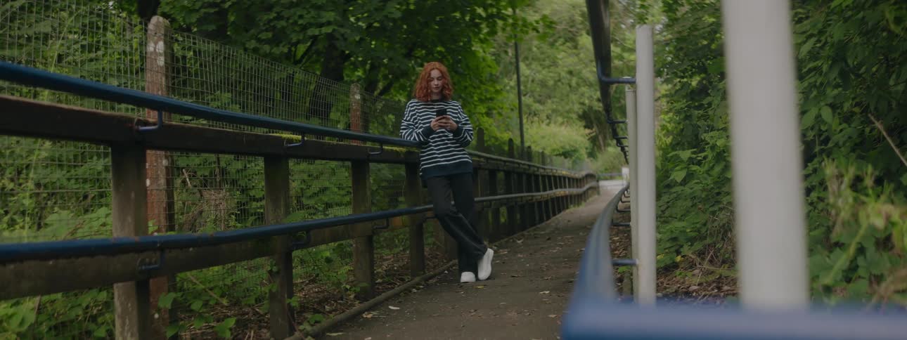 A woman in a striped shirt is using her cell phone while leaning on the railing of a wooden bridge in the forest