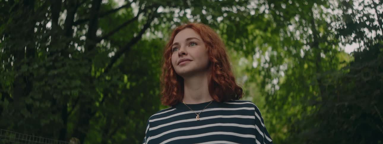 A redhaired woman in a striped shirt with a subtle smile walking in a forest with trees with green leaves