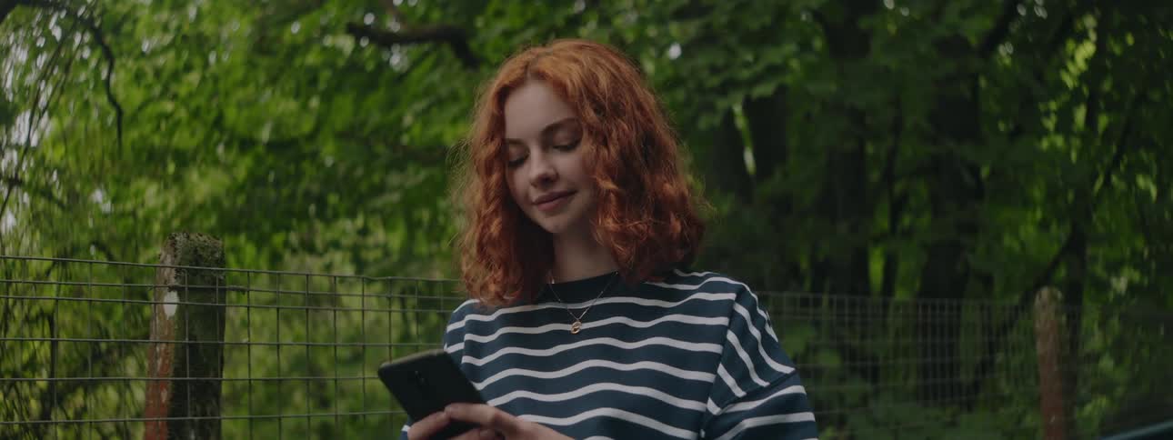 Redhead woman in an outdoor space with trees happily interacting with a smartphone