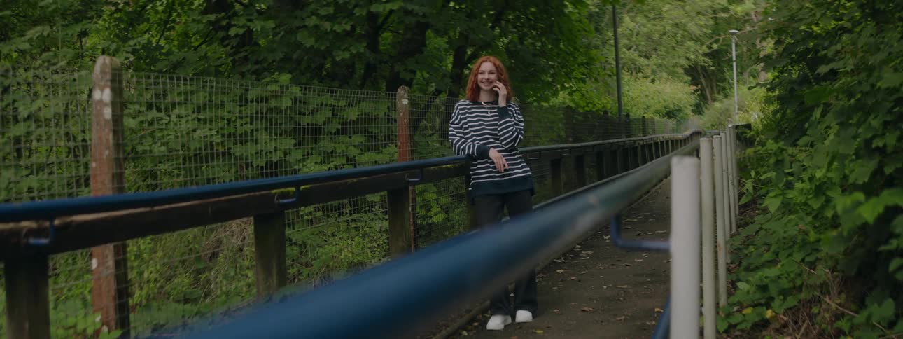 Young redhead woman talking on the phone while standing on a bridge surrounded by trees