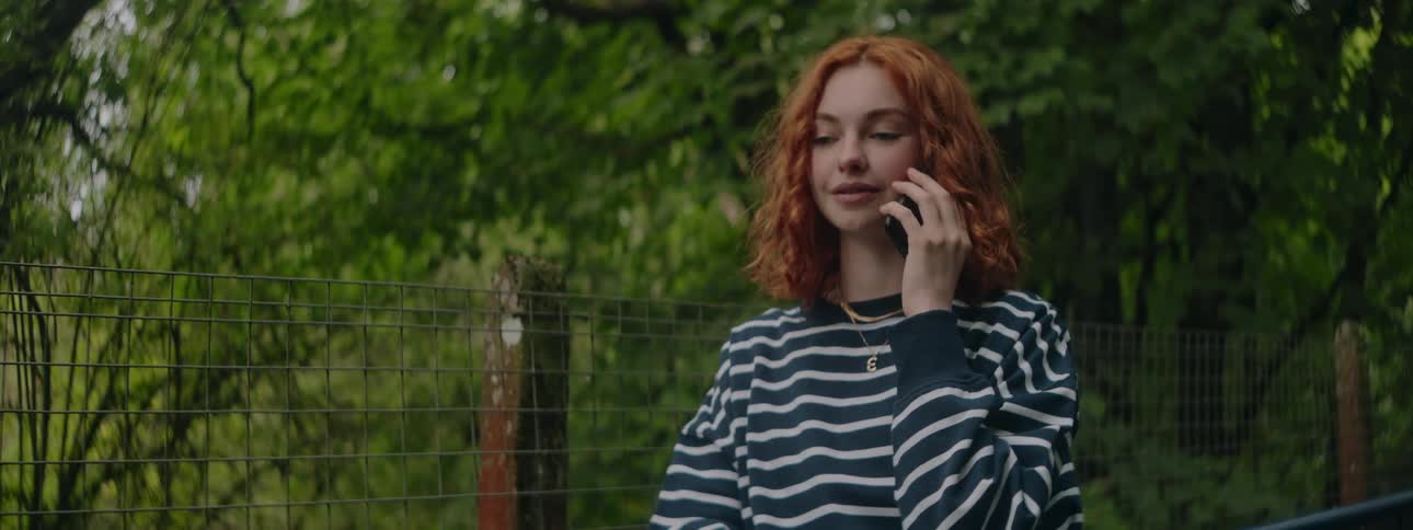 A woman with curly hair stands on a bridge wearing a striped shirt talking on a smartphone