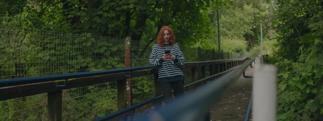 A redheaded teenage girl stands on a foot bridge while texting over a smartphone