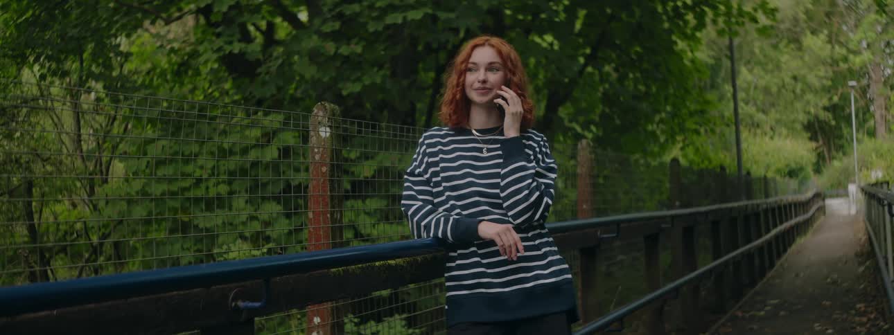 Young redhead woman in an outdoor setting surrounded by trees talking on the phone