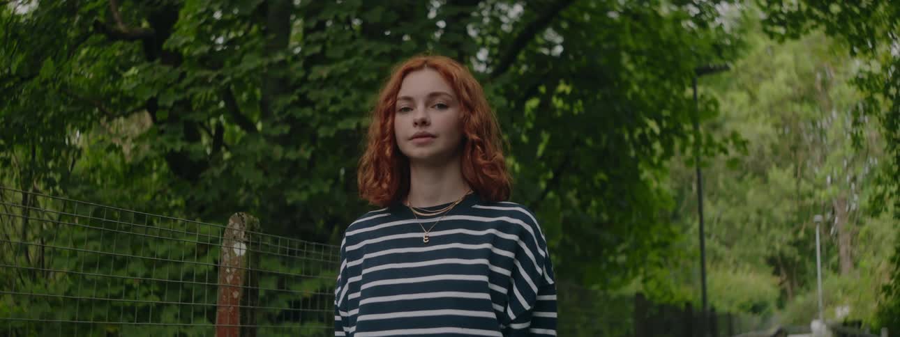 A teenage girl with red hair and a striped shirt stands on a bridge in a forest