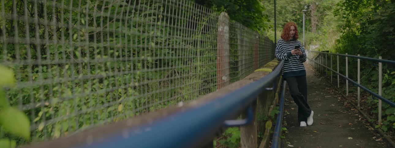 A redhaired teenage girl leans against a bridge railing while she uses her smartphone