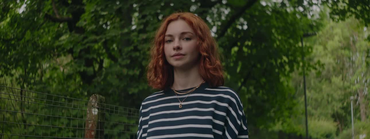 A red headed young woman stands in a tree park and smiles
