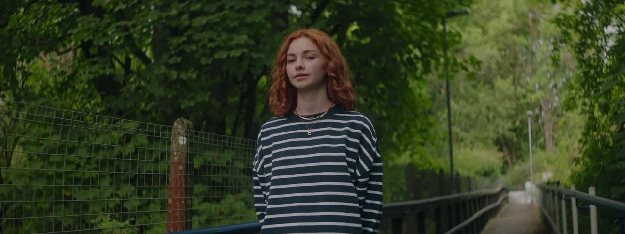 A woman with curly hair stands on a bridge wearing a striped shirt with green foliage in the background