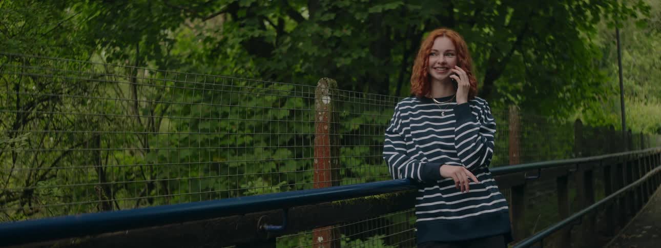 A woman with curly hair stands on a bridge wearing a striped shirt with green foliage in the background