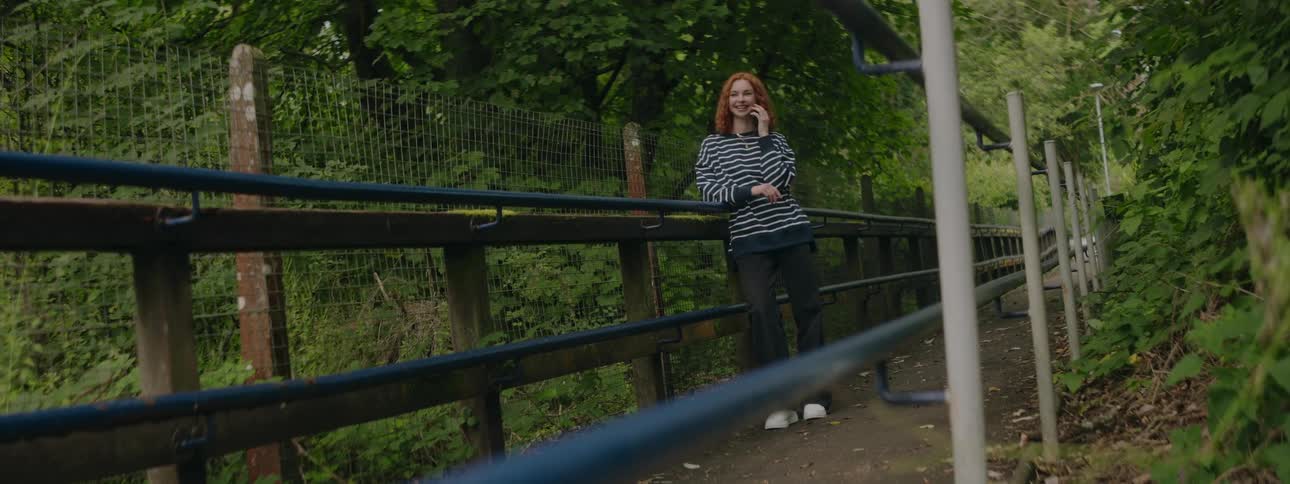 Redheaded woman leans against the rail on a footbridge through the woods and talks on the phone