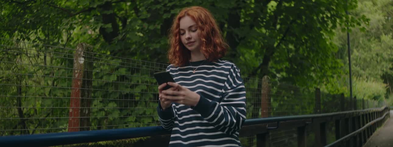A redhaired woman in a striped shirt is typing on her cell phone while leaning on a railing of a bridge and behind her there are some trees
