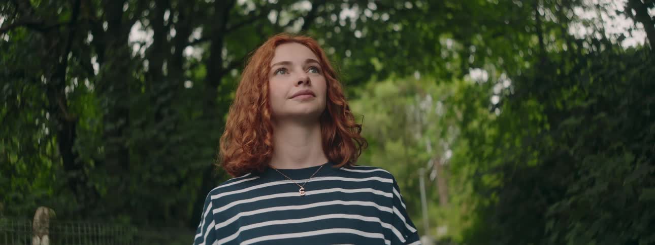A red haired girl smiles and looks about her as she walks through a forest