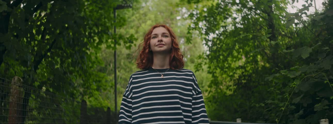 Young redhead woman with happy facial expression walking through a park full of green trees