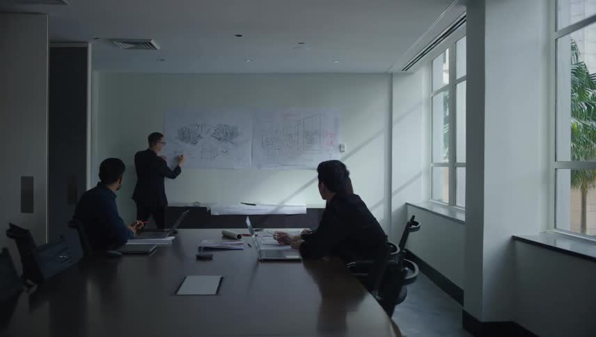 People sitting around a large table in a meeting room watching someone present blueprints on the wall
