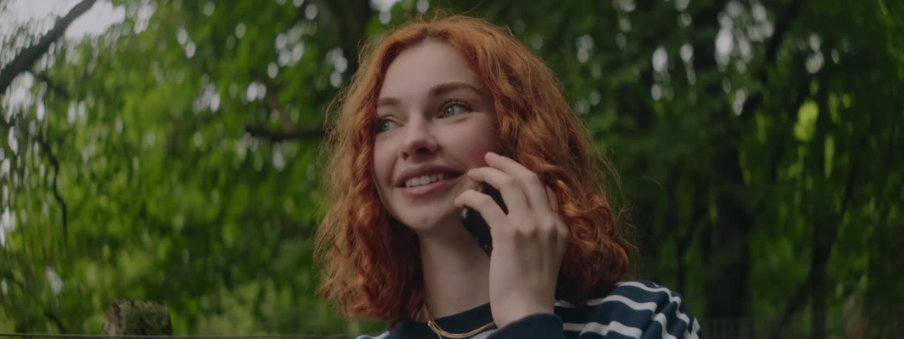 A red haired teenage girl smiles and talks on a smartphone outdoors in a forest