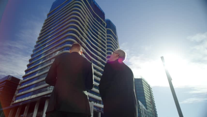 A man standing outside a skyscraper with another man looks up and points