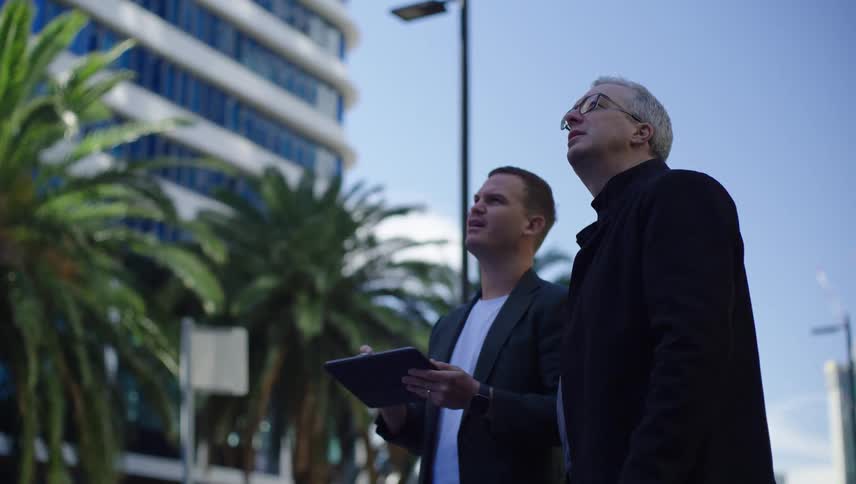 Two men one with a tablet talking in an outdoor area surrounded by buildings