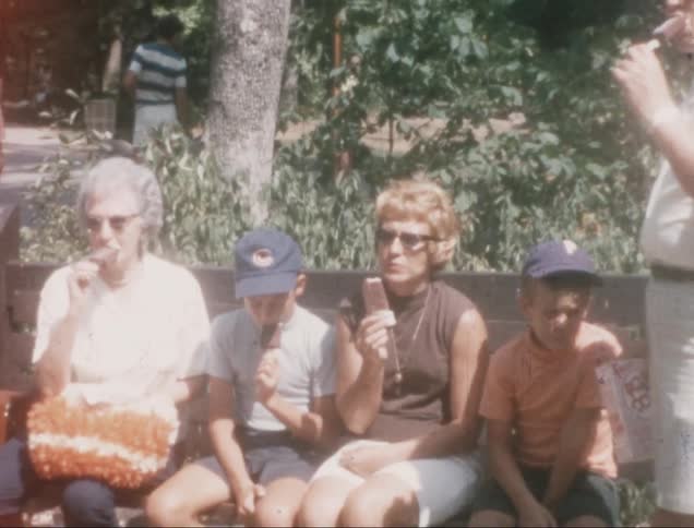 A group of people including an older woman and children are sitting on a park bench eating ice cream
