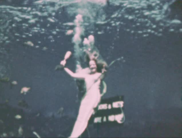 Woman pretending to be a mermaid in an aquarium swings a bell and uses an underwater breathing tube