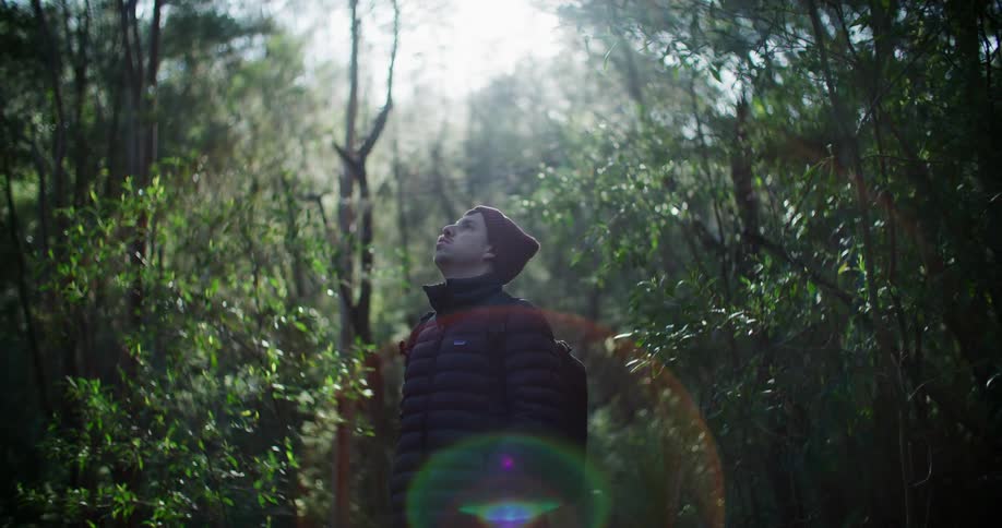 A man in a heavy coat and cap stands in a forest and breathes in the air