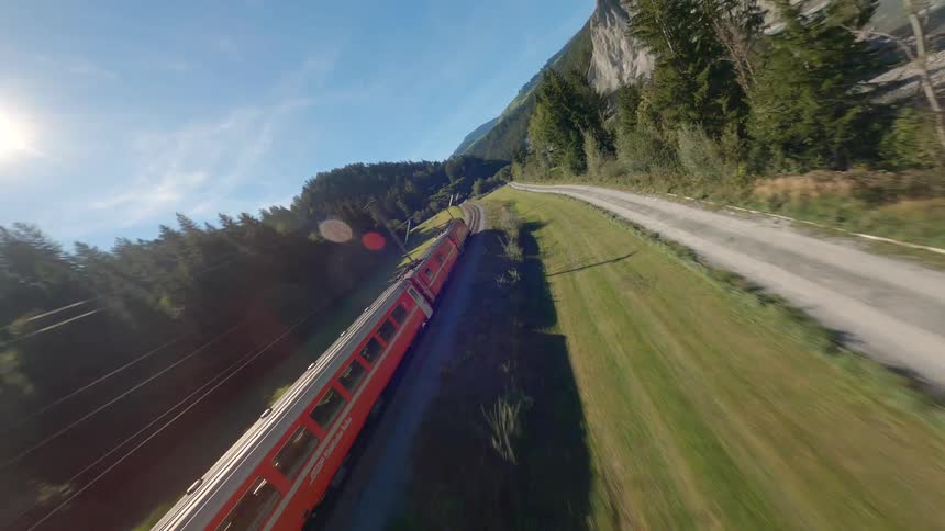 Drone flies towards a Swiss train traveling through an environment surrounded by mountains and vegetation